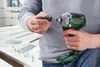 A person is holding a Bosch cordless impact driver and a socket adapter, with construction materials and fasteners on a white surface in the background. Náhľad obrázku produktu