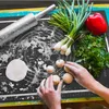 A person's hands are shown working with dough on a black baking mat, with ingredients like green onions, parsley, and a red bell pepper nearby, along with a rolling pin. Voir les images du produit