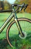 A close-up shot of a green gravel bike's front fork and handlebars, set against a blurred outdoor background of grass and trees. Produktbild-Vorschau