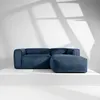 A blue corduroy sectional sofa with an ottoman is displayed on a white platform against a white wall with shadows from window blinds. Produktbild-Vorschau