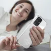 A woman is lying in bed, smiling and holding a transparent phone case with a Samsung Galaxy A51 phone inside. Produktbild-Vorschau