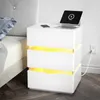 A white nightstand with LED lighting is placed next to a bed, with a laptop and phone charging on top. Náhľad obrázku produktu