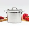 A stainless steel pot with a glass lid sits on a white surface, with a piece of raw meat and some vegetables in the background. Náhled obrázku produktu
