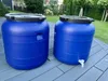 Two blue plastic water barrels are placed on a gray deck in a backyard setting. Produktbild-Vorschau