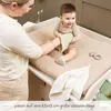 A mother is changing her baby's diaper on a changing table. The baby is smiling and holding a washcloth. Produktbild-Vorschau