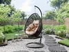 A hanging egg chair with beige cushions sits in a garden setting with a stone pathway and lush greenery. Náhled obrázku produktu