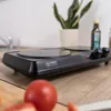 A close-up shot of a black electric double burner on a wooden countertop, with a tomato in the foreground and a plant and bottle in the background. Náhľad obrázku produktu