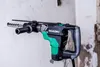 A Hikoki rotary hammer drill is shown mounted on a wall, with a drill bit inserted into the wall. Produktbild-Vorschau