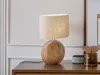 A table lamp with a wooden base and a fabric shade sits on a wooden table. Produktbild-Vorschau