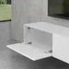 A white TV lowboard with one door open, showing its interior and hinges. The background is a concrete wall and floor, with a window in the distance. Produktbild-Vorschau