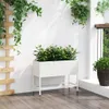 A white planter box filled with green plants sits on a wooden deck against a white wall, with hanging plants and a wooden trellis. Produktbild-Vorschau