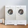 Two white washing machines are stacked on a white pedestal with drawers, in a room with a light gray wall and a patterned rug. Produktbild-Vorschau