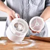 A person is holding a white flour sifter over a wooden surface with flour scattered around it. A rolling pin is also visible on the surface. Produktbild-Vorschau