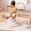 A young girl is playing with a wooden toy toolbox and tools on a rug in a room. Produktbild-Vorschau