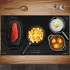 A top-down view of a black induction cooktop with various food items being cooked on it, set against a wooden surface. Produktbild-Vorschau