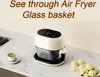 A cream-colored air fryer with a transparent glass basket sits on a marble countertop next to a sink and a cup. A plate of small, round, yellow fruits is also visible. Produktbild-Vorschau