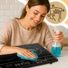 A woman is cleaning an induction cooktop with a spray bottle and a cloth. Voir les images du produit