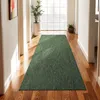 A long green rug is laid out in a hallway with wooden floors. Sunlight streams in from a large window at the end of the hallway, illuminating the rug and the floor. Produktbild-Vorschau