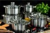 A set of stainless steel pots and pans with glass lids, arranged on a black surface, with some vegetables and spices in the foreground. Náhled obrázku produktu