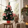 A decorated Christmas tree sits on a wooden stool in a cozy living room setting with a fireplace and armchair in the background. Náhled obrázku produktu
