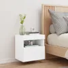 A white floating nightstand is mounted on a white wall next to a bed with a wooden headboard. The nightstand has a plant in a vase, books, and a camera on it. Produktbild-Vorschau