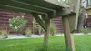 Close-up view of the underside of a wooden garden bench, showing its construction and support legs, set in a grassy garden with a wooden fence in the background. Produktbild-Vorschau