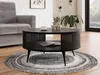 A black round coffee table with a slatted base and a lower shelf is placed on a round rug in a living room setting. A grey sofa is visible to the left, and a large window with greenery is in the background. Náhled obrázku produktu