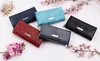 A collection of five women's wallets in different colors, arranged on a white surface with decorative pink flowers and a seashell. Produktbild-Vorschau