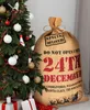 A large burlap sack with Christmas-themed text is placed next to a decorated Christmas tree. Produktbild-Vorschau
