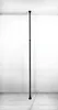 A tall, dark gray pole stands vertically in the center of the frame, anchored to the floor and ceiling. The background is a plain, light gray wall. Náhled obrázku produktu