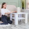 A young woman is sitting on the floor next to a white coffee table, using a laptop. Produktbild-Vorschau