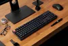 A black mechanical keyboard with RGB lighting is placed on a wooden desk, surrounded by office accessories. Produktbild-Vorschau