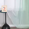 A close-up of a sheer curtain with a green to white gradient, draped over a small table with a framed picture. Produktbild-Vorschau