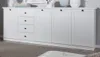A white wooden sideboard with four drawers and four cabinet doors, placed on a carpeted floor. Produktbild-Vorschau