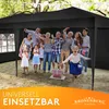 A group of people, including children and adults, are gathered under a black canopy tent, celebrating what appears to be a party or event. Some are holding drinks, and there are gift boxes on the ground. Produktbild-Vorschau