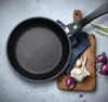 A frying pan is placed on a light gray surface next to a wooden cutting board with onions and garlic. Produktbild-Vorschau