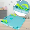 The underside of a teal-colored baby activity table with lime green straps is shown, with a magnified inset view of one of the straps. The background is blurred, showing a nursery setting with a crib and dresser. Podgląd zdjęcia produktu