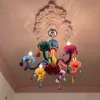 A colorful, whimsical chandelier hangs from a decorative ceiling medallion. Produktbild-Vorschau