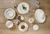 A table setting with various pieces of tableware, including plates, bowls, cups, and cutlery, arranged on a wooden surface. Produktbild-Vorschau