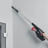 A person is using a cordless vacuum cleaner with an extension wand to clean a corner of a wall. Voir les images du produit