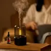 A person is blurred in the background, reading a book, while an aroma diffuser and essential oil bottle are in the foreground. Podgląd zdjęcia produktu