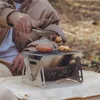 Silberner tragbarer Campinggrill mit einer Wurst und einem Hähnchenflügel, die mit einer Zange in der Hand einer Person im Freien gegart werden Anteprima dell’immagine prodotto