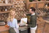A couple in a kitchen, one person is cooking while the other is showing them a painting. Voir les images du produit