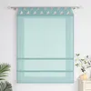 A teal roman blind with a decorative top edge hangs on a curtain rod against a white wall. A green plant is visible on the left, and a small decorative branch with flowers and candles sits on a white cabinet on the right. Produktbild-Vorschau