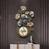 A wall clock with a decorative metal frame shaped like ginkgo leaves, mounted on a dark gray wall above a cabinet. Produktbild-Vorschau