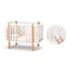 A white baby crib with wooden accents, presented on a white background. A circular inset shows a close-up of a hand holding a white component of the crib. Produktbild-Vorschau