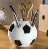A soccer ball shaped pen holder on a wooden desk with various writing utensils and a globe in the background. Produktbild-Vorschau