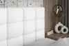 A close-up view of a white upholstered headboard of a bed, with a bedside table and decorative items in the background. Produktbild-Vorschau