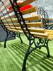 A close-up view of a park bench with wooden slats and black metal legs, placed on artificial grass. Produktbild-Vorschau