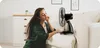 A woman is sitting on the floor next to a couch, enjoying the breeze from a fan. Anteprima dell’immagine prodotto